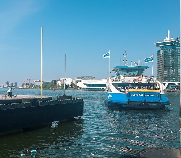 A blue ferry labeled IJVEER 54 AMSTERDAM sails on the water near a pier where a cyclist rides with modern buildings in the background