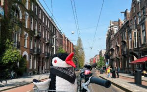 Miško the knitted penguin wearing a bandana sits in a bike basket on a street in Amsterdam with brick buildings and tram lines