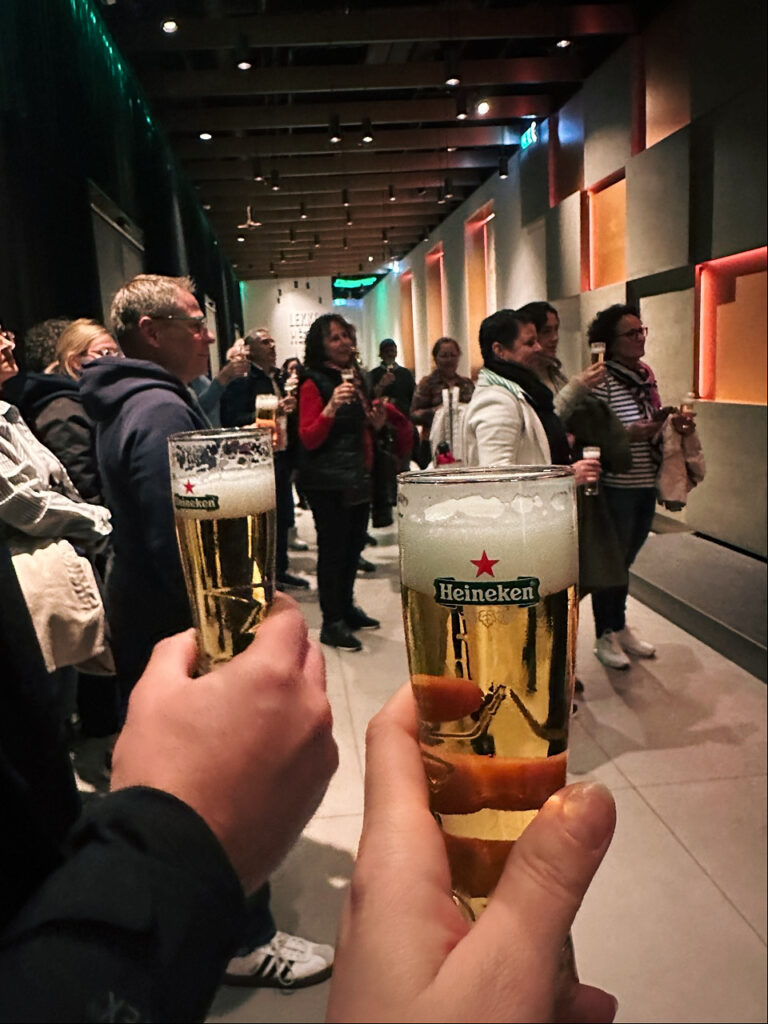 Two hands holding Heineken beer glasses in front of a crowd of visitors tasting beer at the Heineken Experience Amsterdam