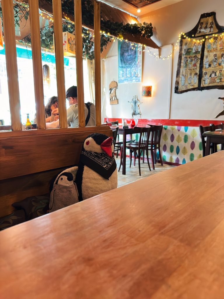Miško the penguin wearing a black bandana sits at a wooden table in Lalibela Amsterdam with diners in the background