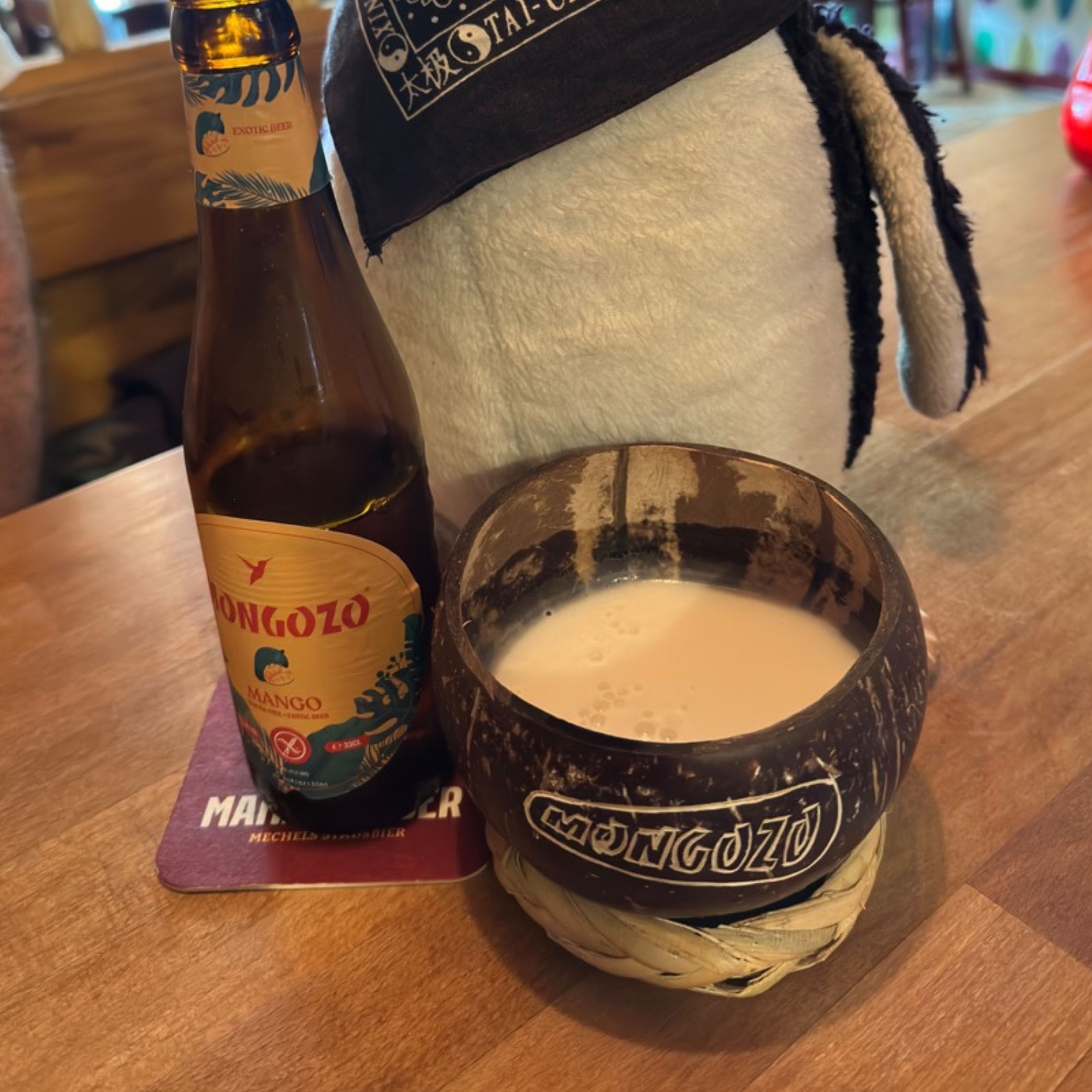 Mongozo Mango beer bottle and a coconut cup with a creamy drink sit on a wooden table in front of the penguin Miško