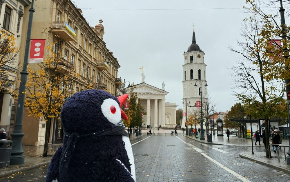 Miško the knitted penguin looks towards a white cathedral and a tall bell tower in Vilnius on a cloudy day