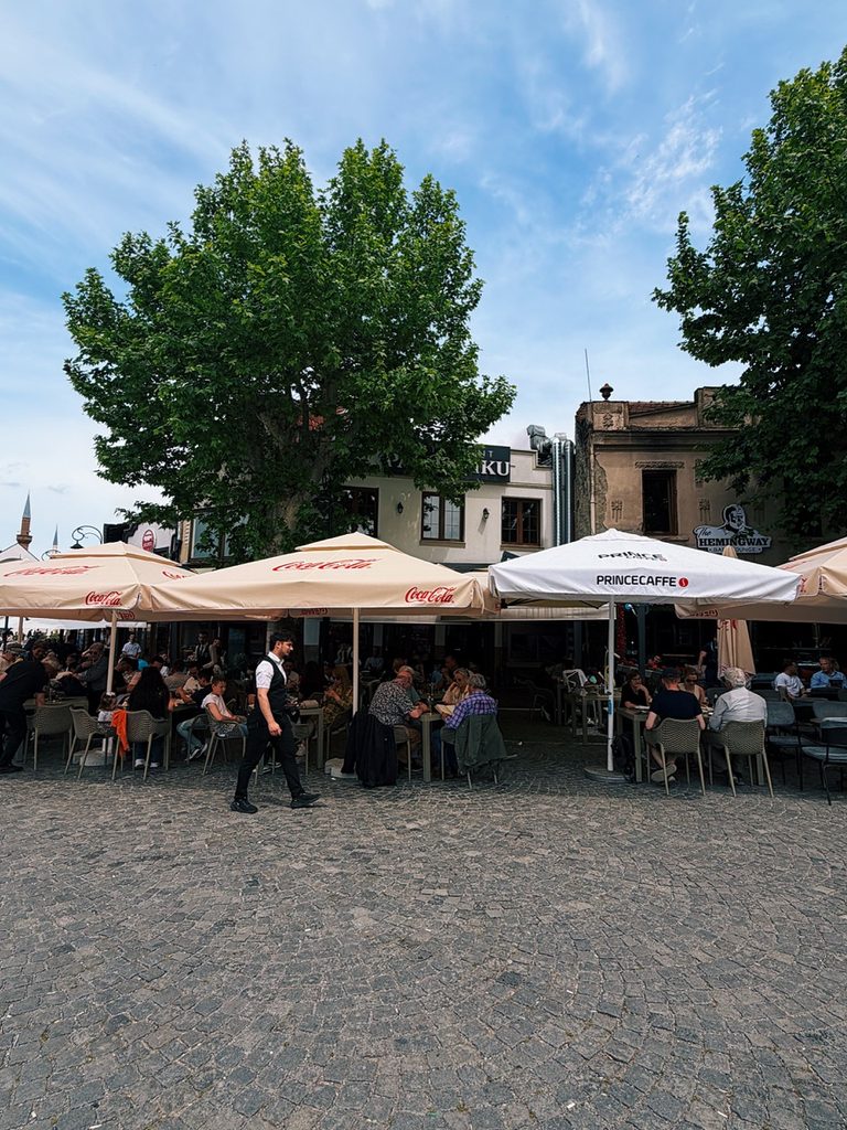 Diners sit under Coca-Cola and Prince Caffe umbrellas outside Restaurant Familjar Pashtriku Prizren on a cobblestone square