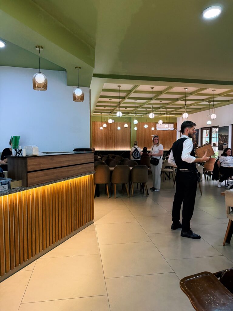 Waiter holding a menu stands in the dining room of Restaurant Familjar Pashtriku near a wooden counter with vertical slats