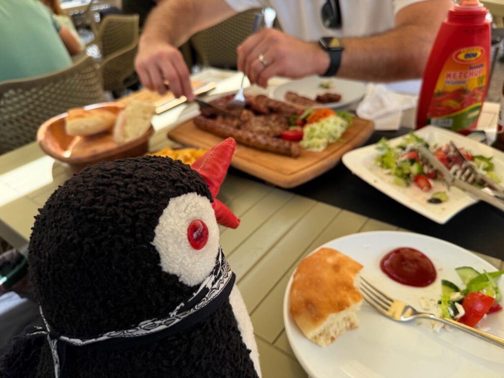 Miško the knitted penguin sits in front of a table with grilled meats, bread, and salad at Restaurant Familjar Pashtriku Prizren