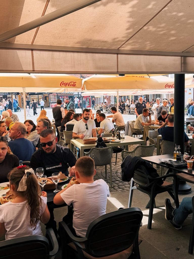 Diners sit at tables under large Coca-Cola umbrellas on the sunny outdoor terrace of Restaurant Familjar Pashtriku Prizren