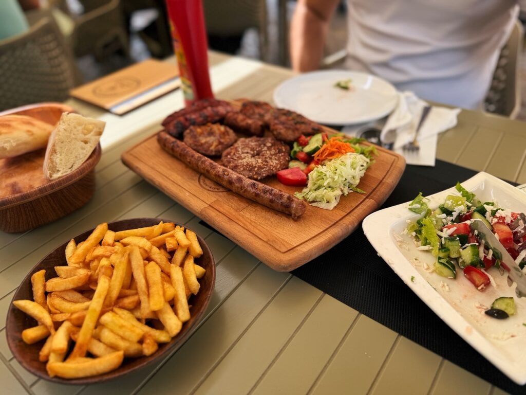 A wooden board of mixed grilled meats and salad sits beside french fries and bread at Restaurant Familjar Pashtriku Prizren