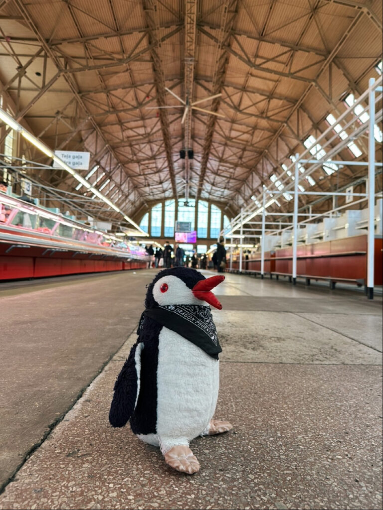A knitted penguin toy perched on a market stall with the sign jedi Teja gaja ANIZESZNE in the background