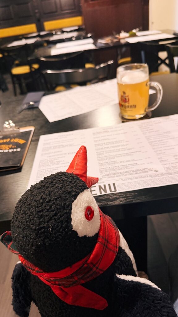 Miško the penguin looks at a menu on a table with a glass of beer in the background at Smoke House Košice