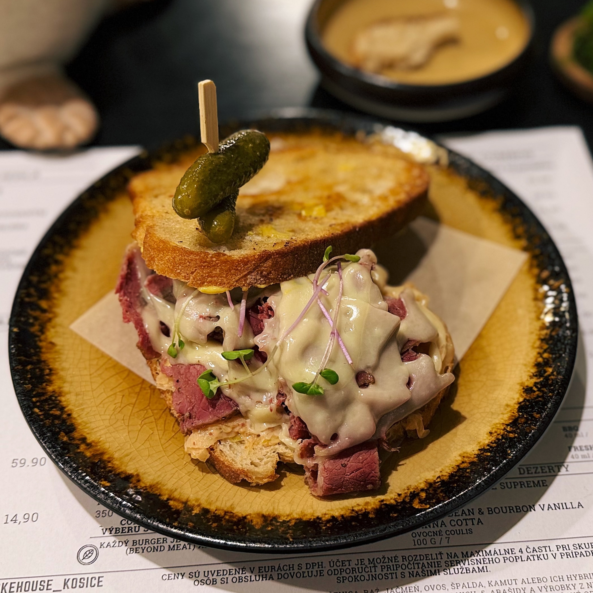 Pastrami sandwich with creamy sauce and microgreens topped with pickles on a skewer on a plate at Smoke House Košice