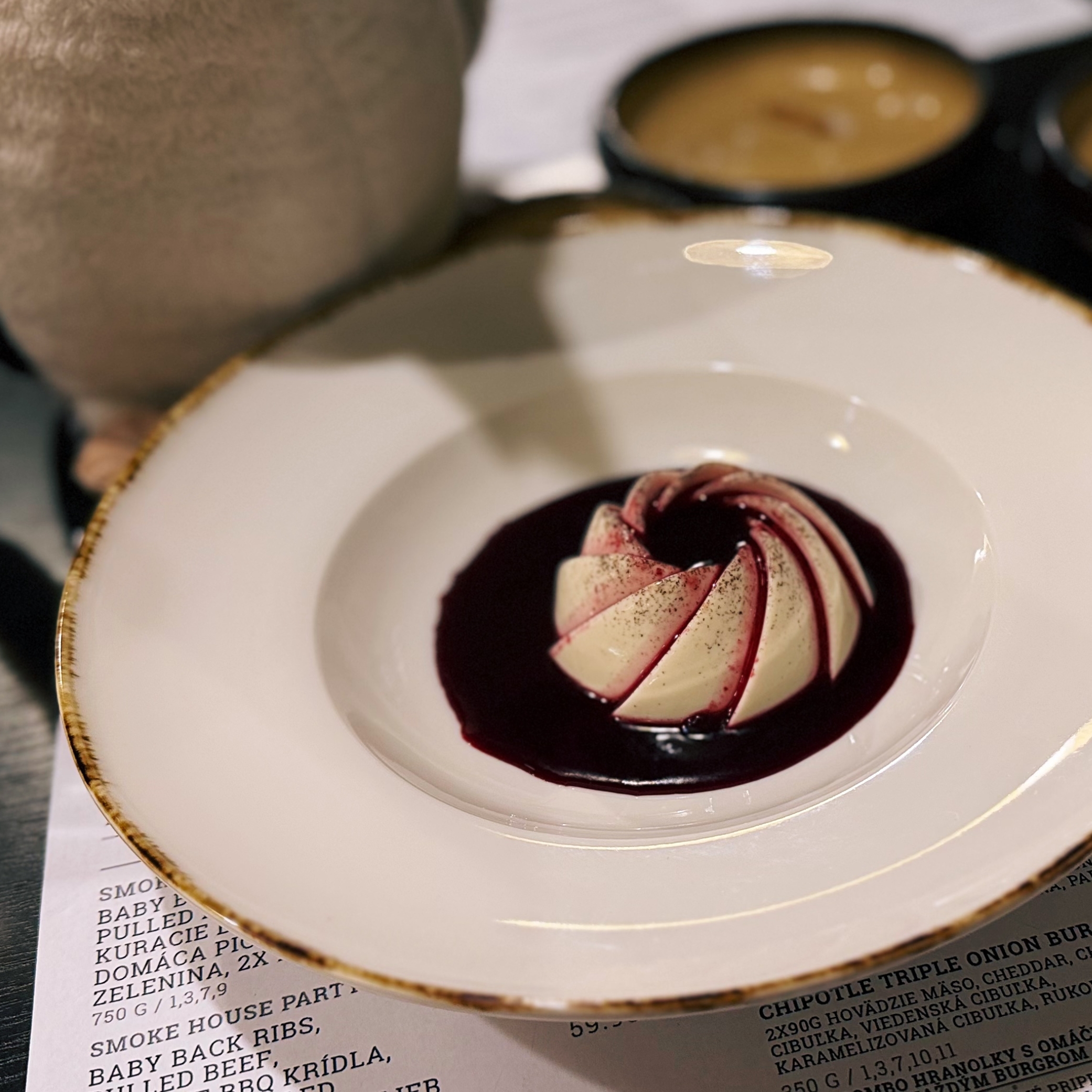 Sliced creamy dessert with dark berry sauce on a white plate with a gold rim resting on a menu at Smoke House Košice