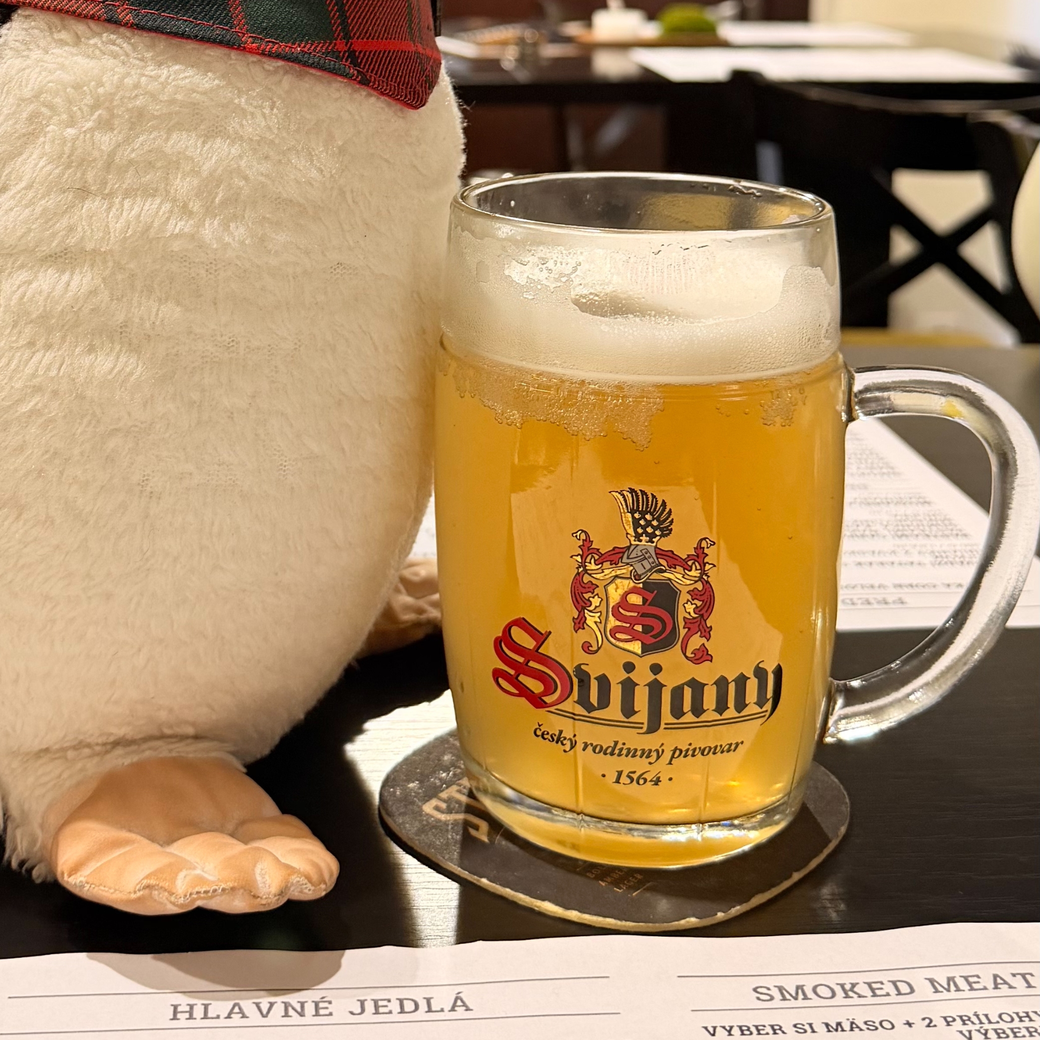 Miško the penguin sits next to a mug of Svijany beer on a table at Smoke House Košice above a menu