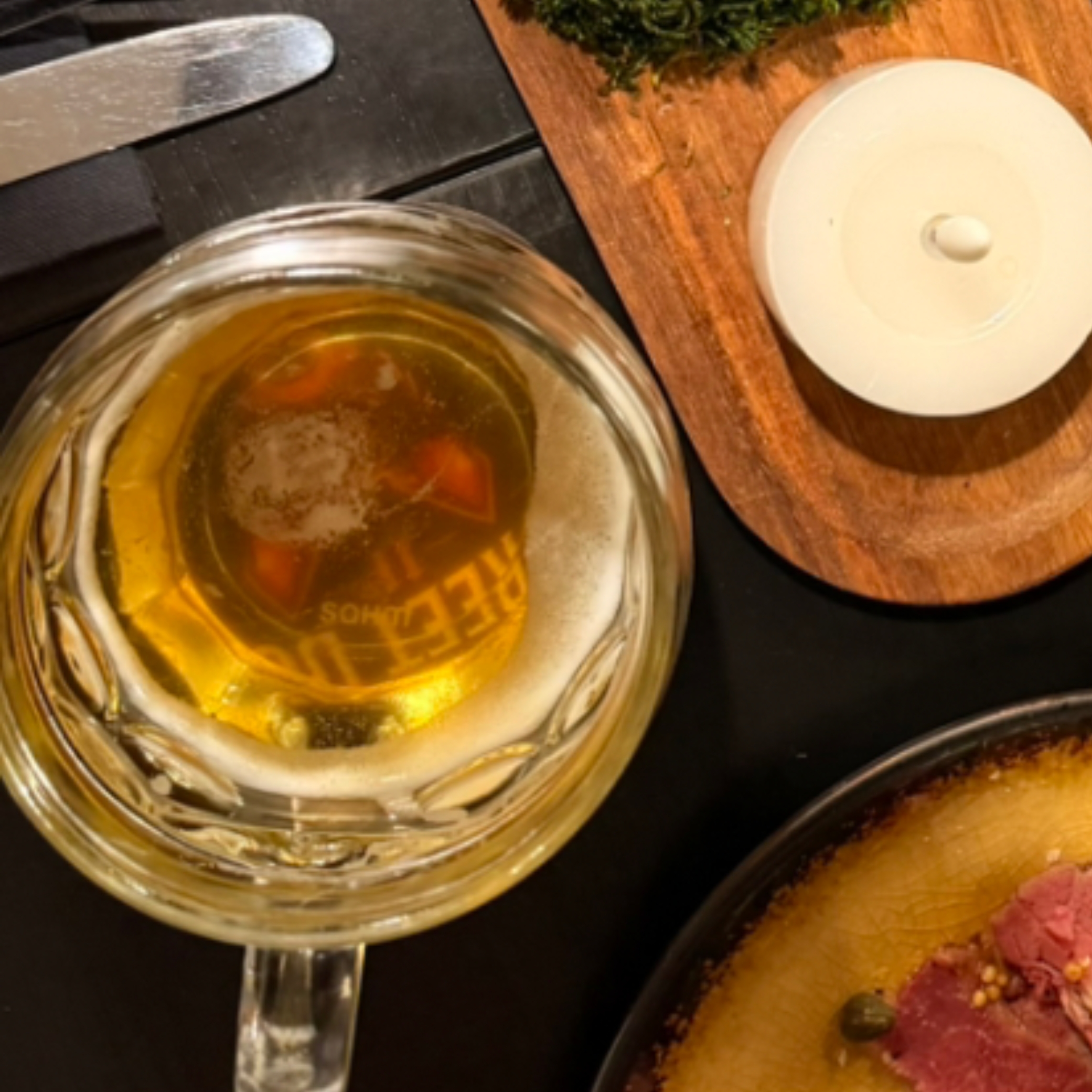 A glass mug of beer sits next to a wooden board with a white ceramic lid and a plate of sliced meat at Smoke House Košice