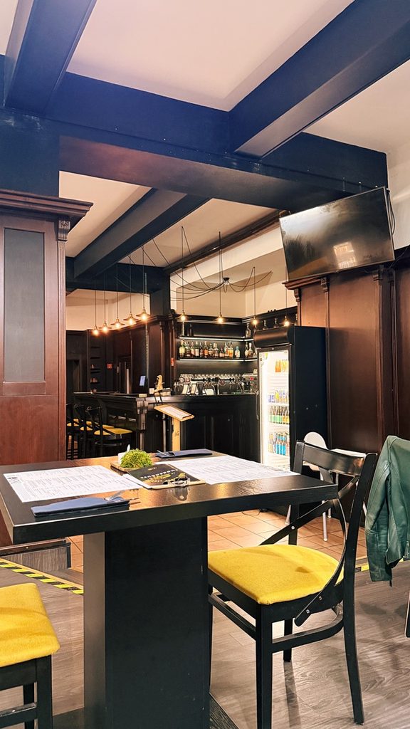 Interior of Smoke House Košice with a table, yellow-cushioned chairs, dark beams, a TV, and a bar area stocked with bottles