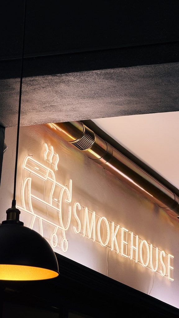 A white neon sign on a wall features a barbecue smoker graphic next to the word SMOKEHOUSE beneath a metal ventilation pipe