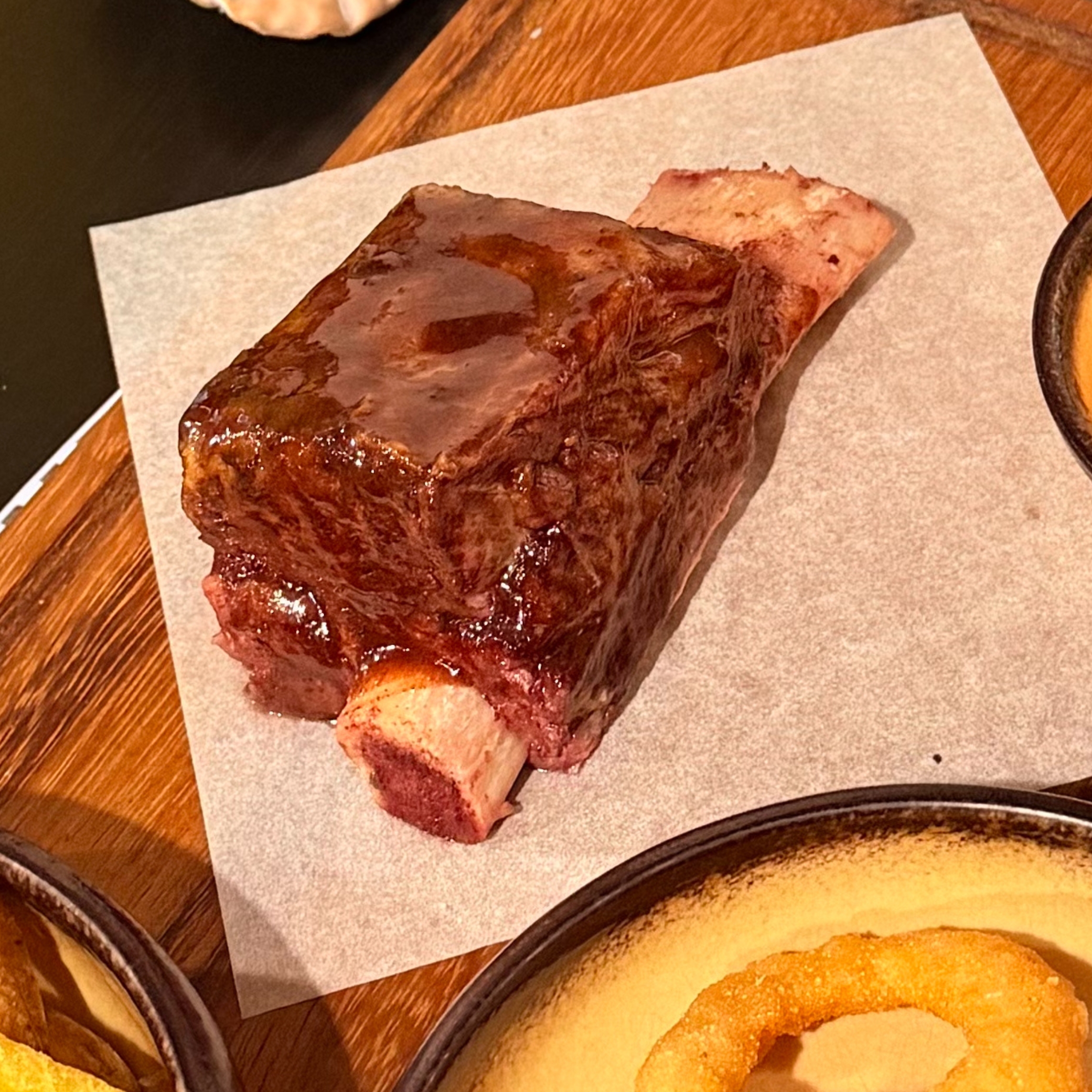 A large glazed rib with a bone resting on parchment paper on a wooden table near a bowl of onion rings