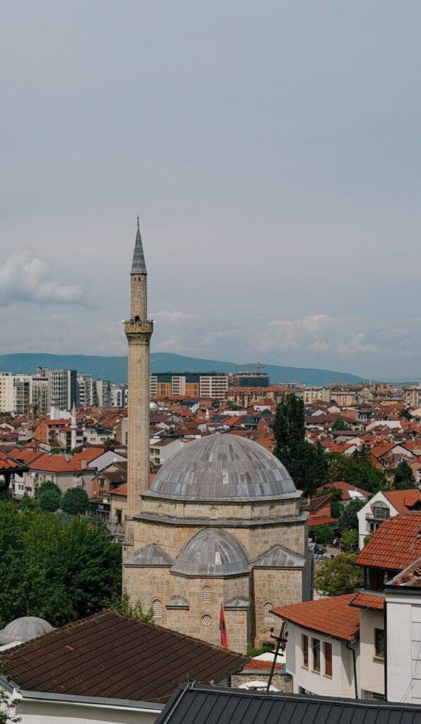 A stone mosque with a tall minaret and grey dome stands among the red rooftops of Prizren with mountains in the distance