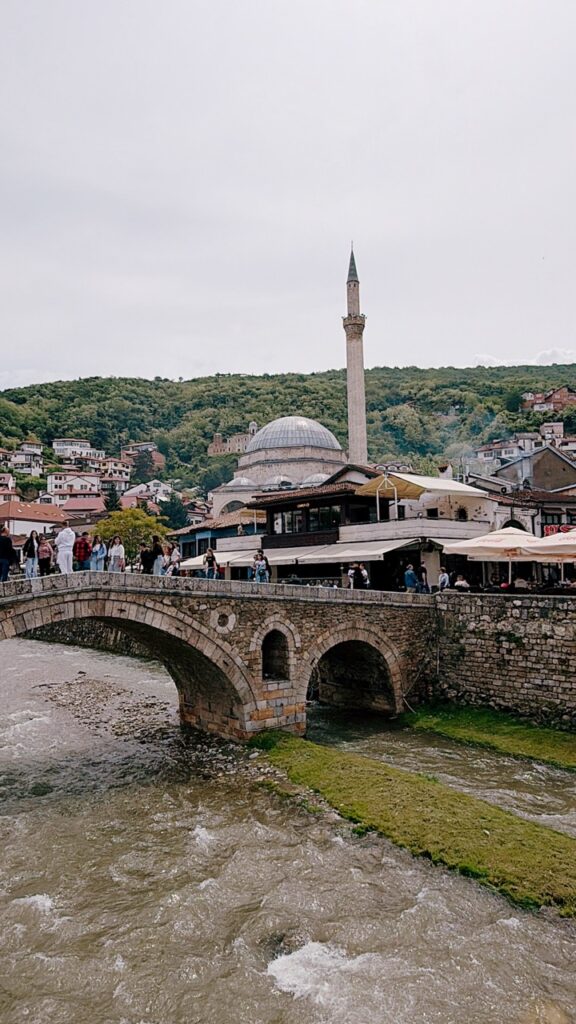 People walking across a historic stone bridge over a river in Prizren with a mosque and tall minaret in the background