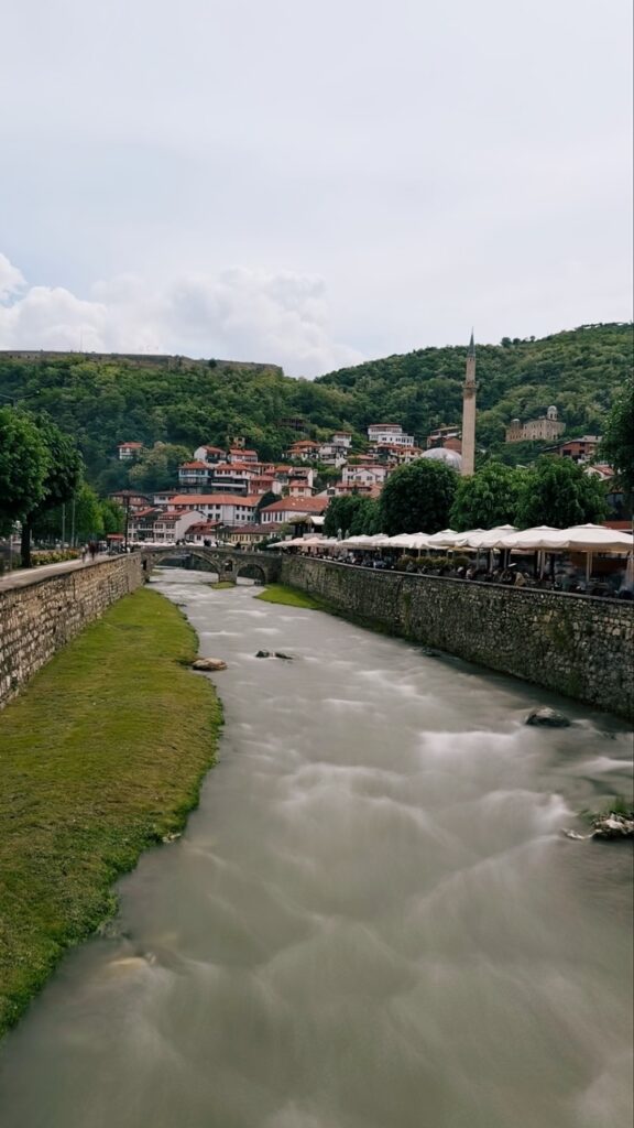 A river flows through Prizren past stone walls and white umbrellas towards a green hillside town with a tall minaret