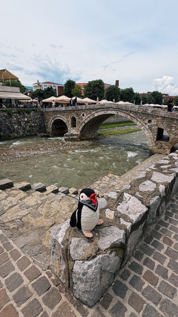 Miško the knitted penguin sits on a stone wall overlooking a river and an old stone bridge in Prizren
