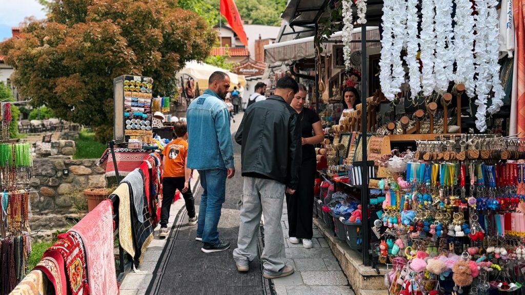 People browse a souvenir market in Prizren with colorful keychains and hanging white decorations near a railing with textiles