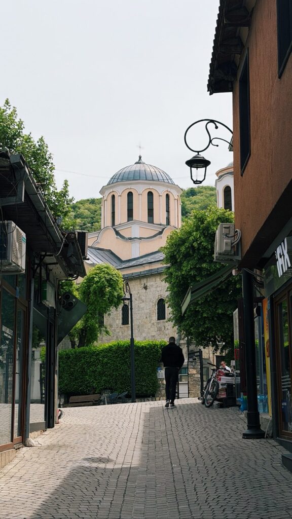 A man walks down a cobblestone street in Prizren towards a large church with a dome framed by buildings and trees