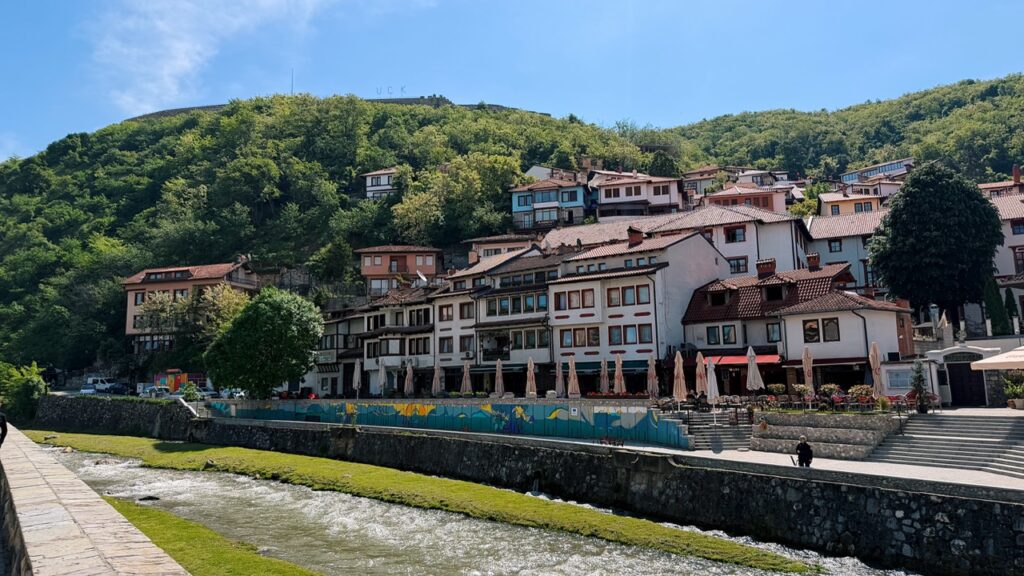 Traditional houses with red roofs line the riverbank in Prizren, backed by a steep green hill under a blue sky