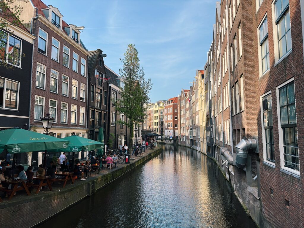 A canal in the Netherlands lined with tall brick buildings and an outdoor cafe with green umbrellas where people are sitting