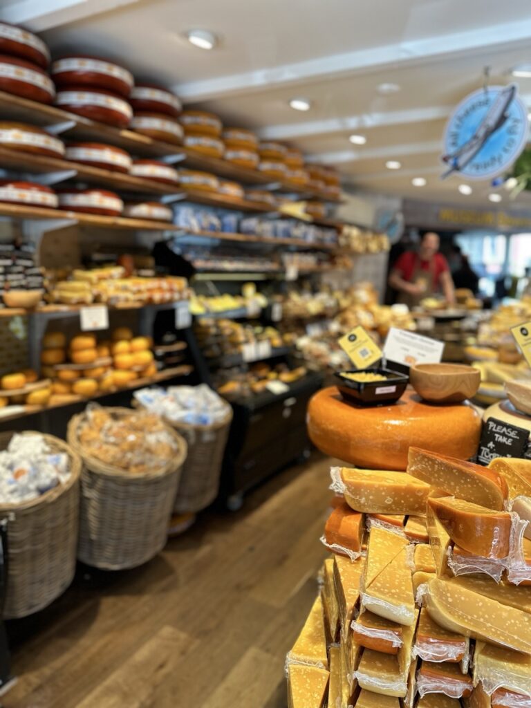 Shelves of large cheese wheels and baskets in a shop with a stack of sliced cheese wedges and a sign reading Please Take A Ticket