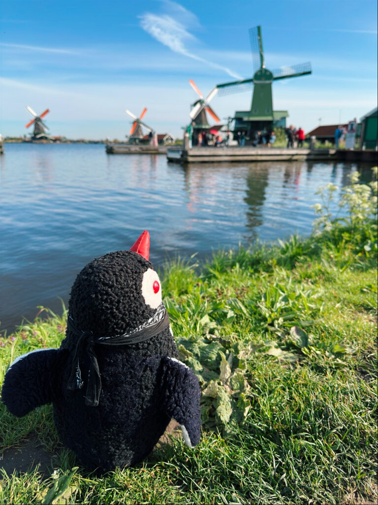 Miško the knitted penguin sits on the grass looking at windmills across the water in the Netherlands