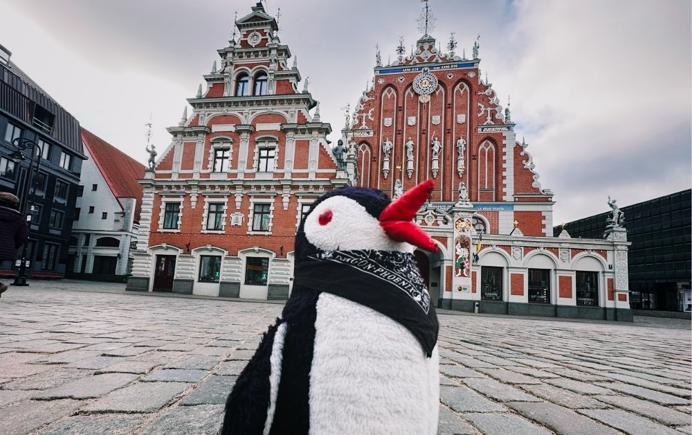 Miško the penguin wearing a black bandana stands on a cobblestone square in Riga with ornate red buildings behind