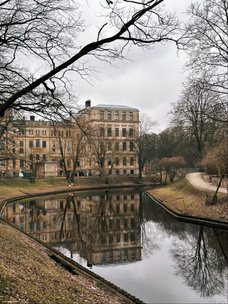 A grand beige building with rows of windows reflects in a calm canal in Riga, framed by bare tree branches