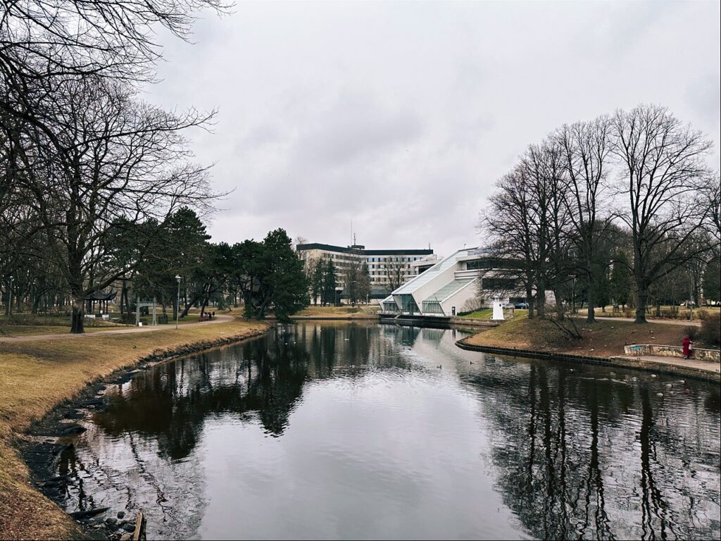 Bare trees line a calm river in Riga reflecting a modern white building with a sloping roof on the opposite bank