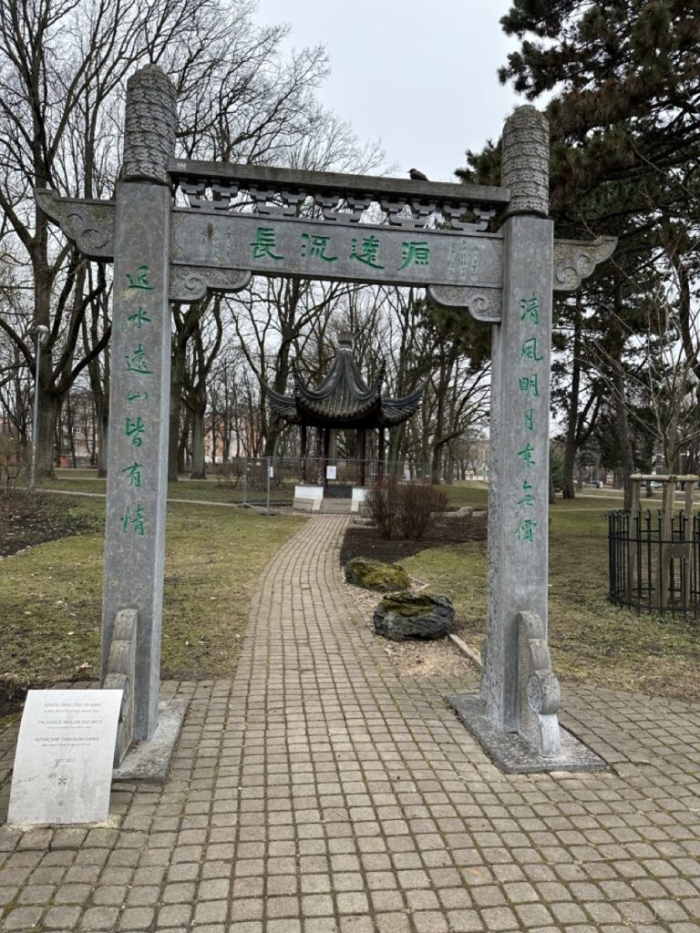 A stone Chinese gate with green characters frames a brick path leading to a traditional pavilion in Riga Latvia