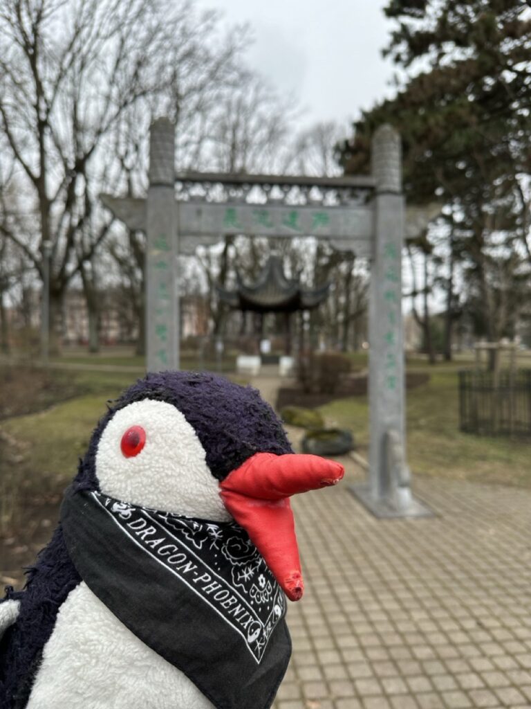 Miško the knitted penguin wearing a black Dragon-Phoenix bandana stands in front of a Chinese archway in Riga