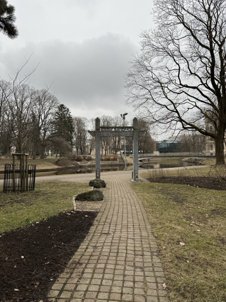 A paved path leads through a park in Riga, Latvia towards a wooden gate near a canal surrounded by bare trees