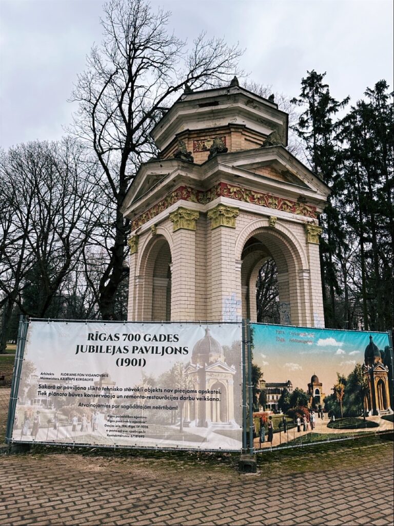 A stone pavilion with arches stands behind a fence with a banner reading RĪGAS 700 GADES JUBILEJAS PAVILJONS (1901)