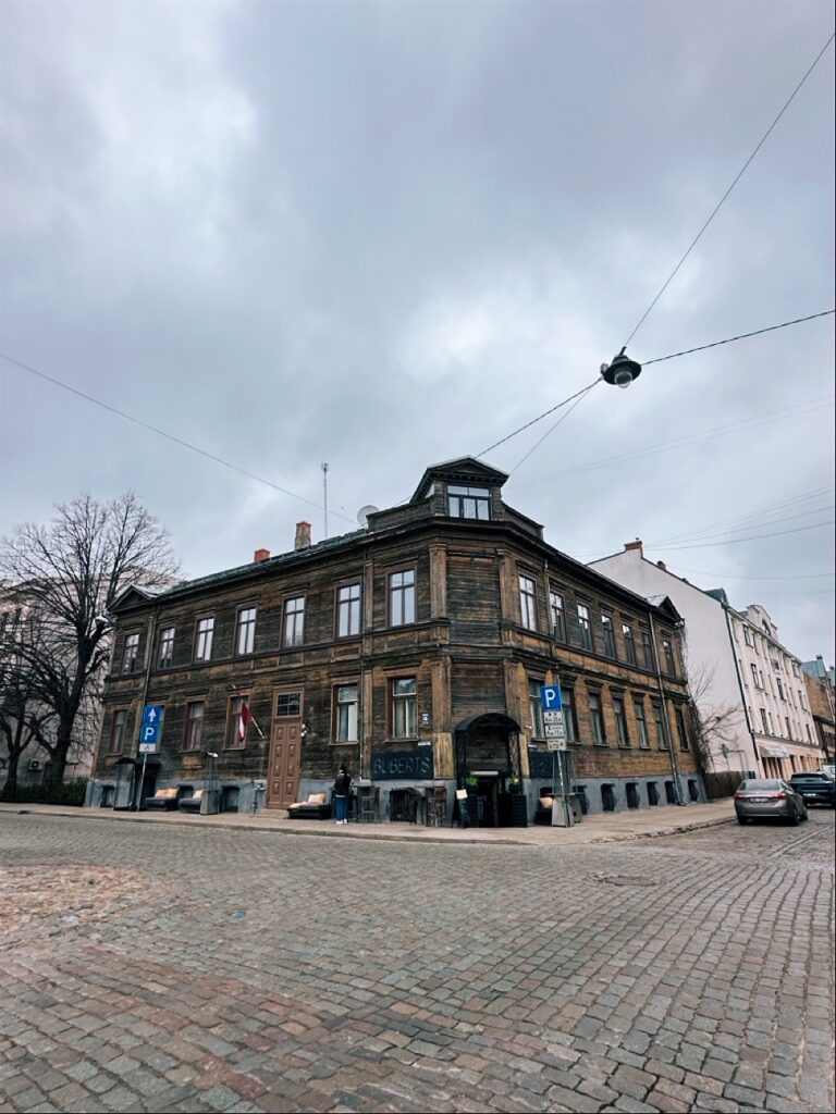 A dark wooden corner building with a sign reading ALBERTS on a cobblestone street in Riga