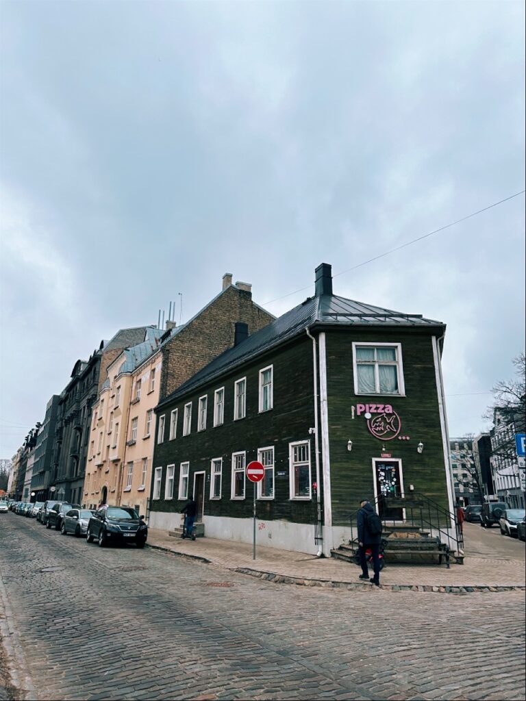 Dark green wooden corner building with a Pizza sign on a cobblestone street in Riga, Latvia lined with parked cars and a pedestrian