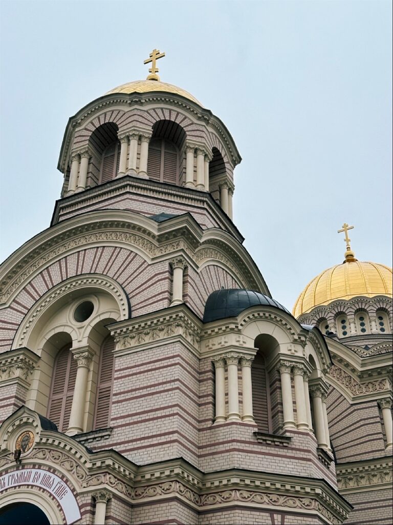 Ornate cathedral in Riga featuring pink and white striped brickwork, arched windows, and golden domes topped with crosses