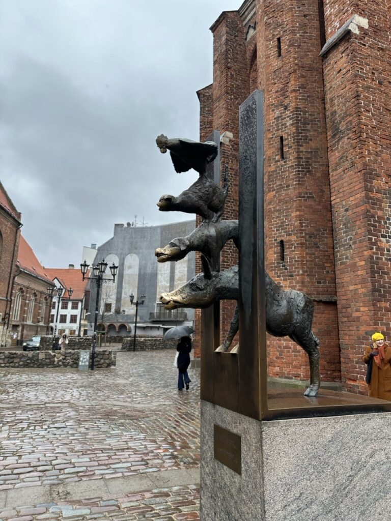 Bronze sculpture of a donkey, dog, cat and rooster stacked vertically in Riga with a brick cathedral in the background