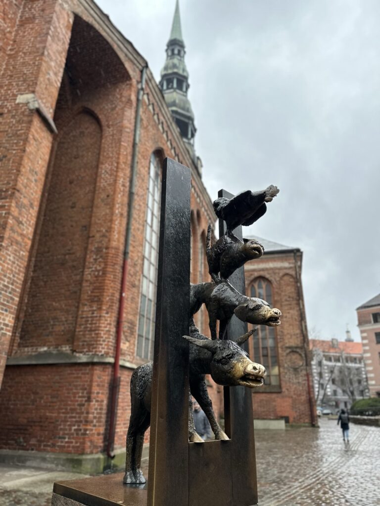 Bronze sculpture of a donkey, dog, cat and rooster stacked vertically in front of a red brick church in Riga