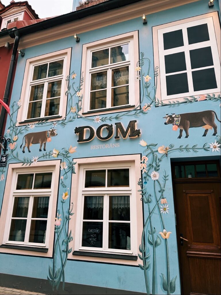 Blue facade of DOM RESTORĀNS in Riga, Latvia featuring painted cows and floral vines around windows advertising hot wine