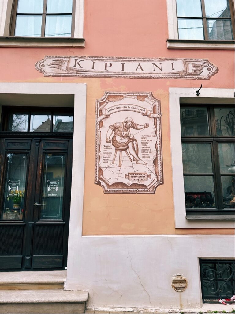 Peach building facade in Riga with a KIPIANI sign above a mural of a shoemaker working next to a dark door