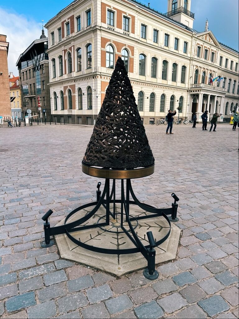 A large metal Christmas tree sculpture stands on a cobblestone square in front of a grand building in Riga Latvia