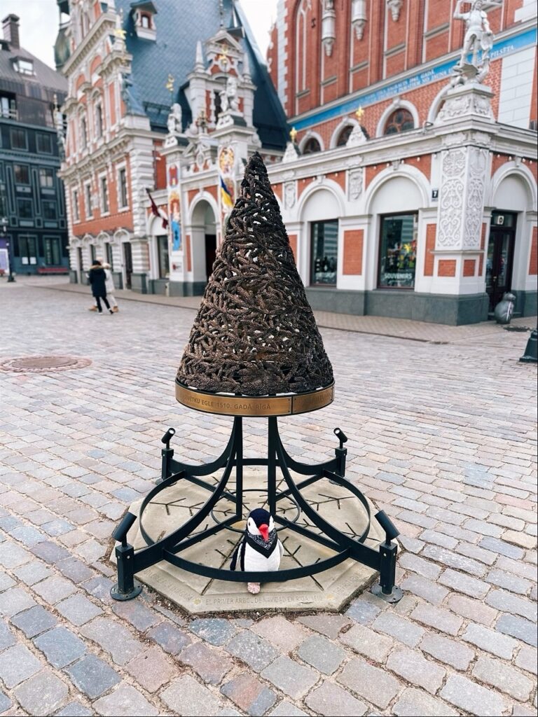 Miško sits at the base of a cone sculpture marking the first Christmas tree in Riga, Latvia with historic buildings behind