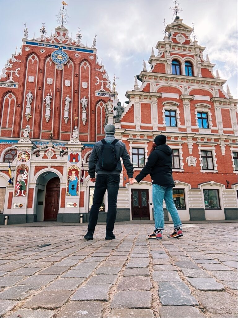 Two travellers hold hands on a cobblestone square facing ornate red brick buildings with white statues and gables in Riga
