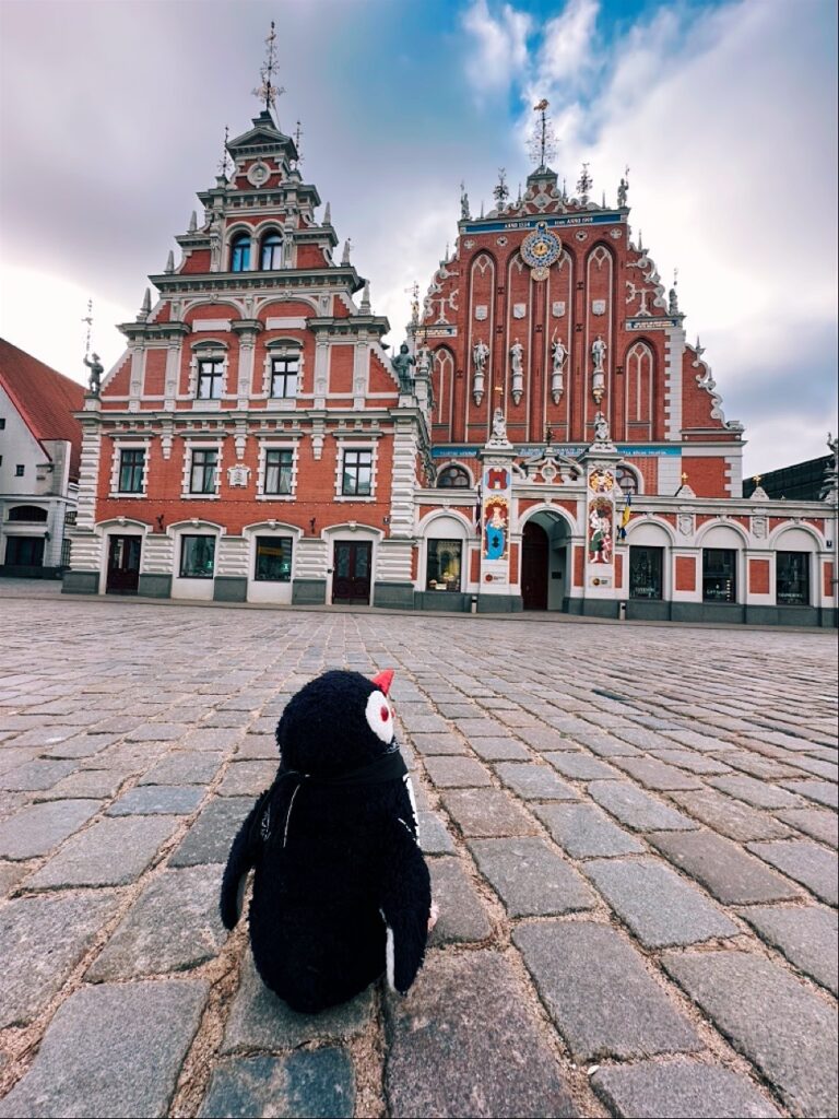 Miško the knitted penguin toy sits on a cobblestone square facing two ornate red buildings in Riga, Latvia