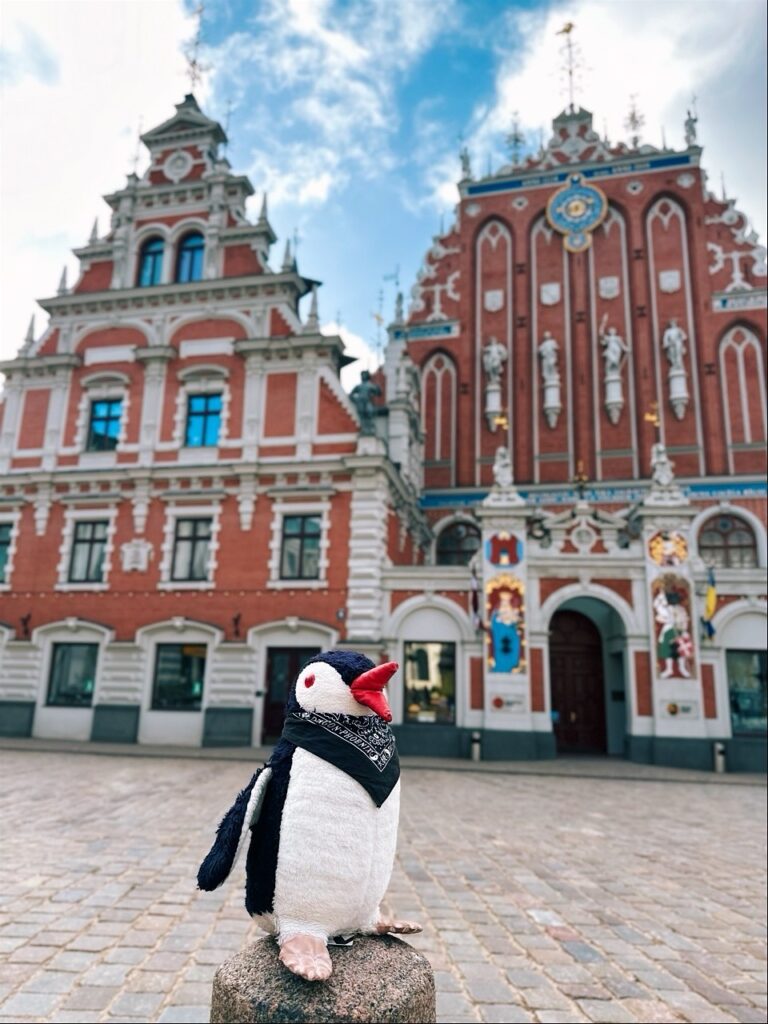 Miško the knitted penguin wearing a black bandana sits on a stone post in front of ornate red buildings in Riga