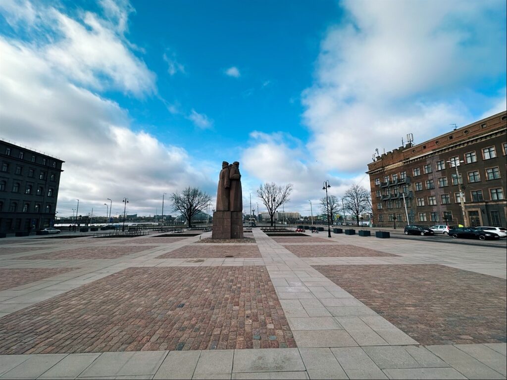 A wide paved square in Riga with a central stone monument of two figures under a blue sky with scattered clouds